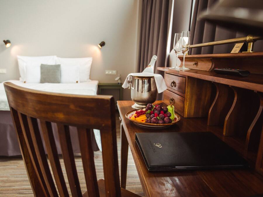 A hotel room with a bed, bedside table with a bottle, glasses, and a fruit plate.