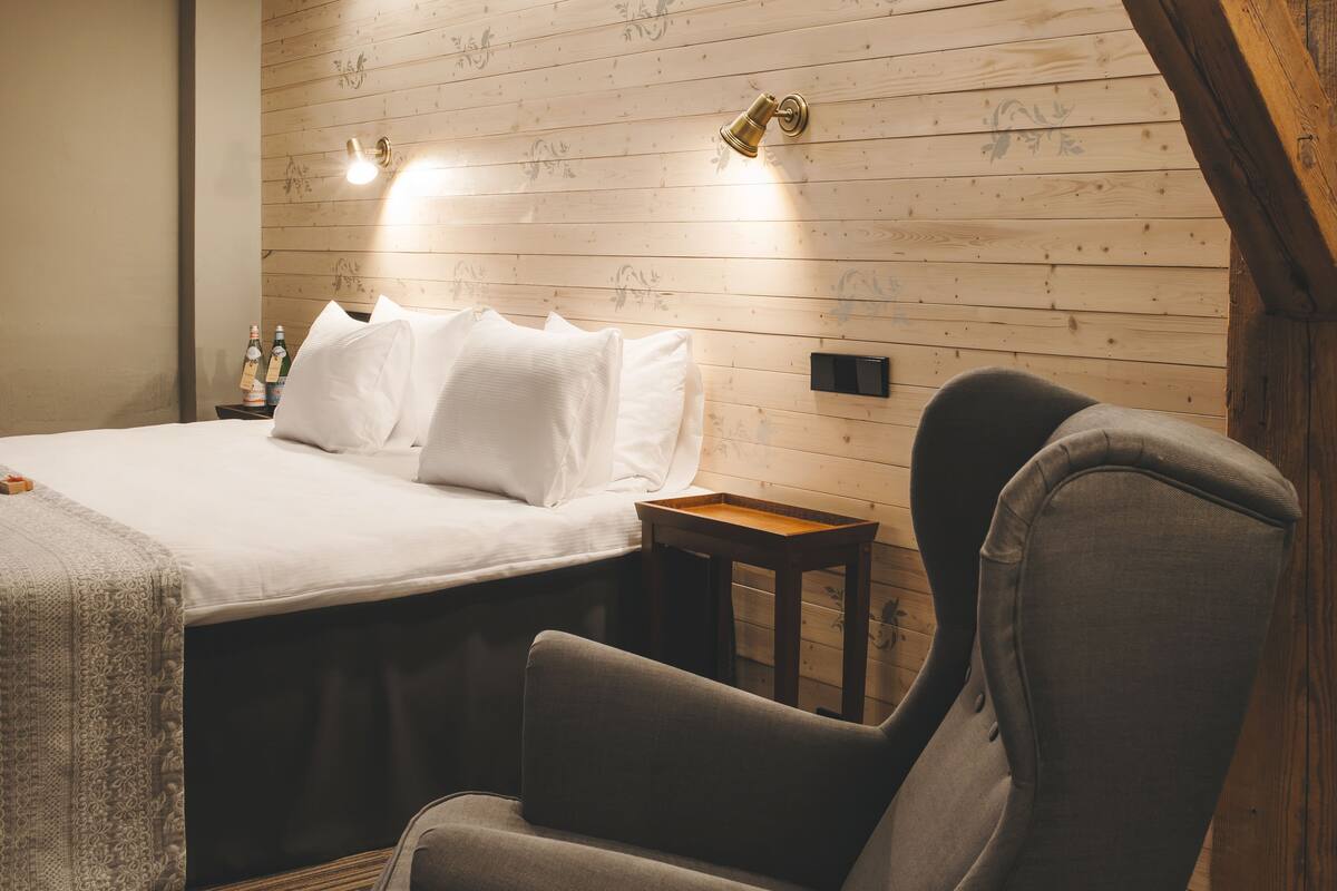 A hotel room with a bed, a chair, a nightstand with bottles, and wooden paneling on the walls.