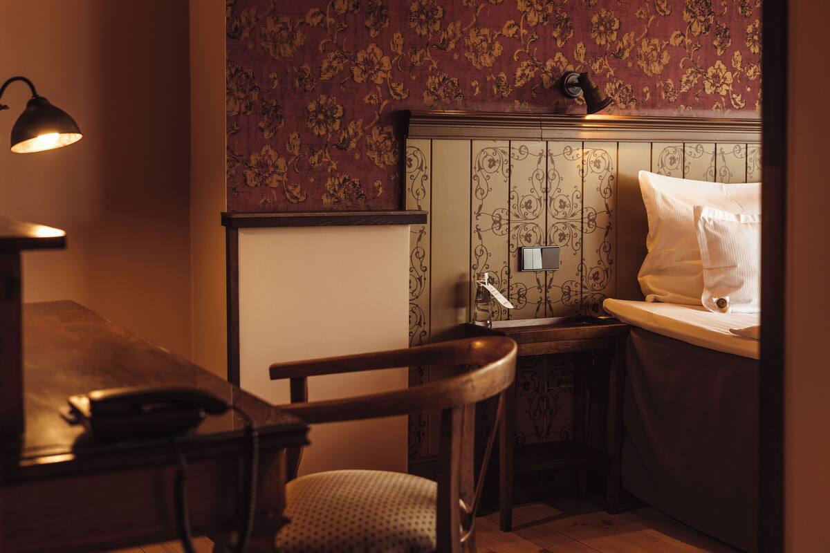A small, dimly lit room with a wooden desk, a chair, a bed with a white pillow, and a wall with floral wallpaper.