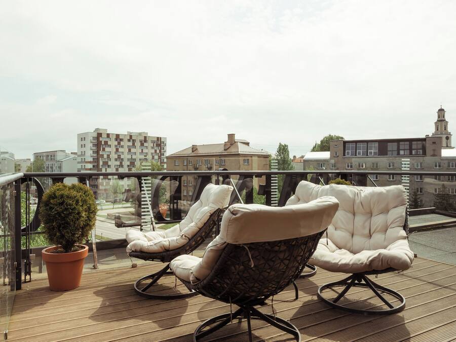 Un balcón con dos sillones acolchados y una planta en maceta, con vistas a un paisaje urbano que incluye edificios y una torre.