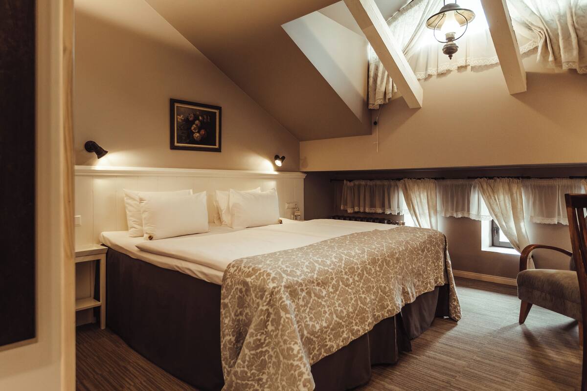 A bedroom with a large bed, a nightstand, a chair, and a window with curtains.