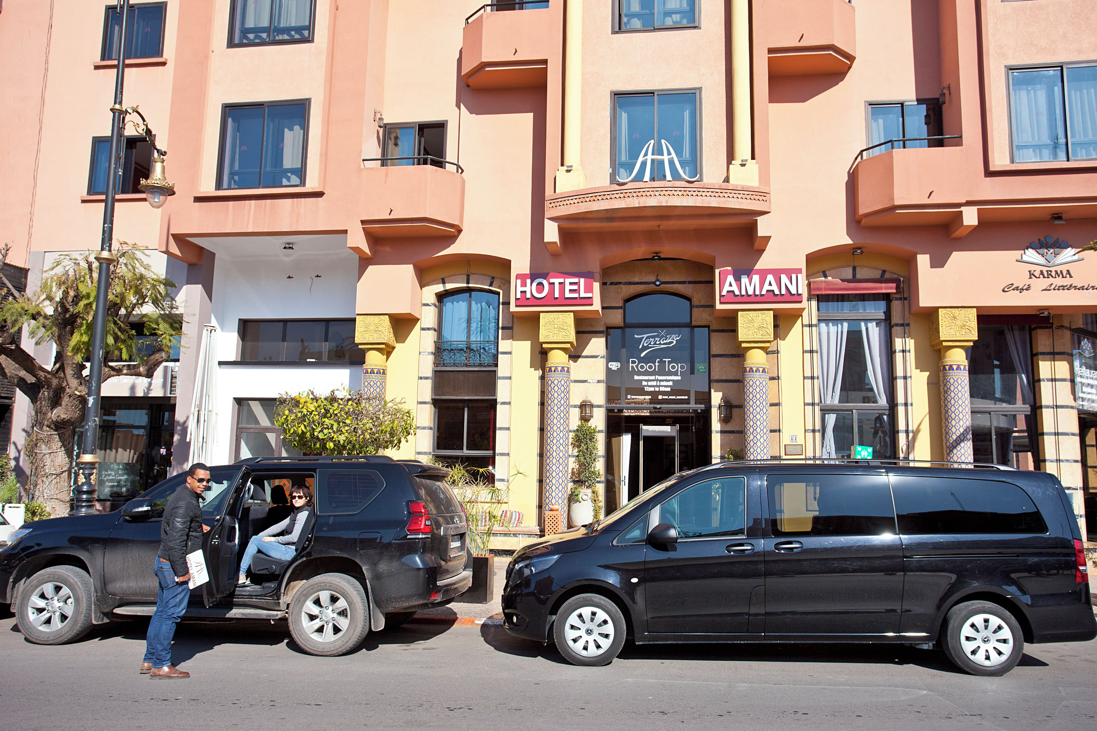 Un hotel con un letrero que dice 'HOTEL' y 'AMANI' en la fachada, un coche estacionado y una persona de pie junto al vehículo.