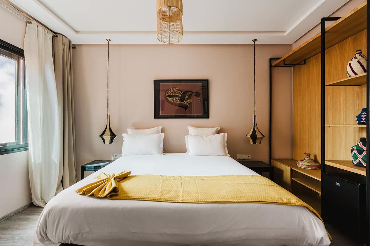Una habitación de hotel con cama, dos mesitas de noche, un armario, una lámpara colgante y un cuadro en la pared.