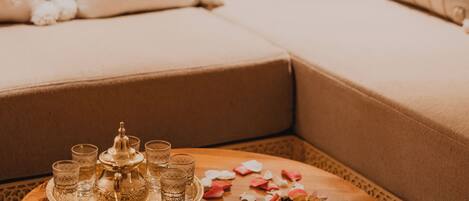 Una habitación acogedora con un sofá decorado con cojines, una mesa de centro de madera con una bandeja de vasos y una tetera, y pétalos de rosa esparcidos.