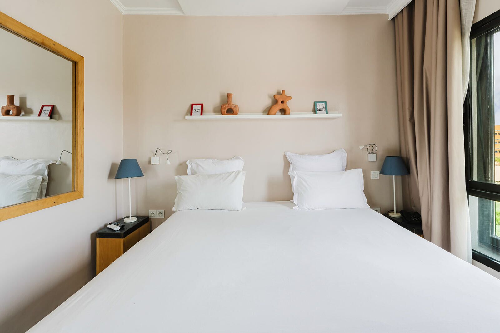 Una habitación de hotel con una cama grande, dos mesitas de noche, un espejo, un estante con adornos y una ventana con cortinas.