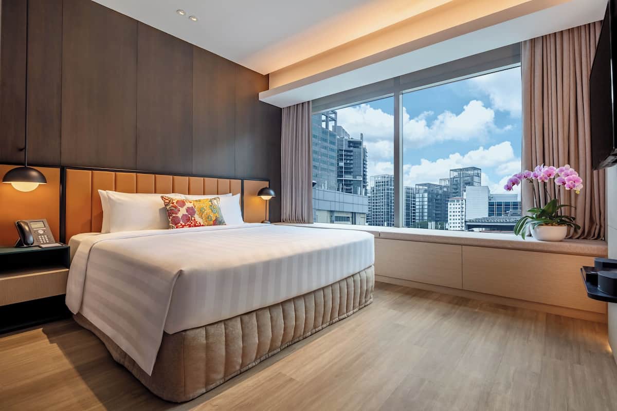 A modern hotel room with a large bed, bedside lamps, a window with a city view, and a small table with a phone.