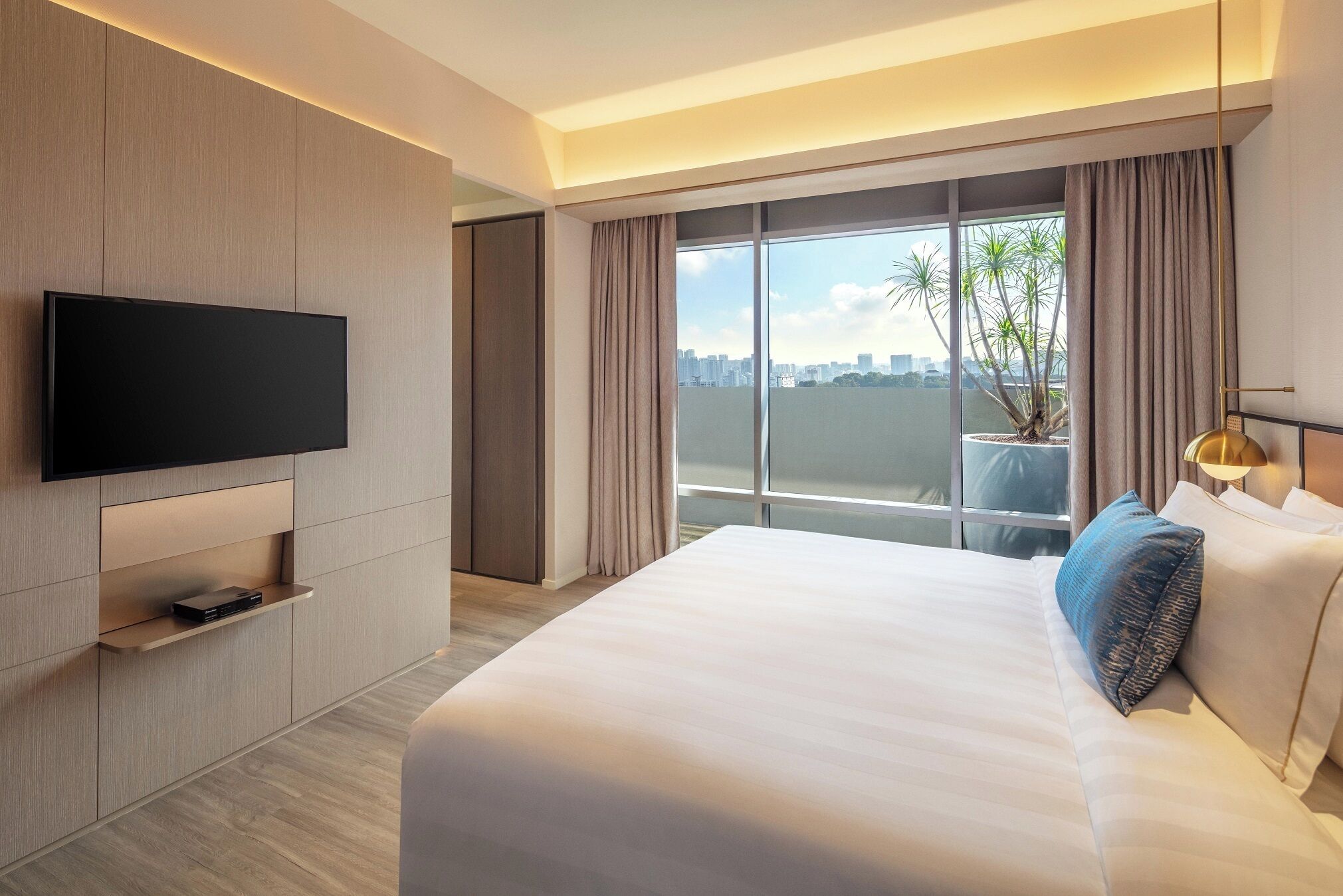 A modern hotel room with a large bed, a flat-screen TV mounted on the wall, a potted plant, and a city view through large windows.
