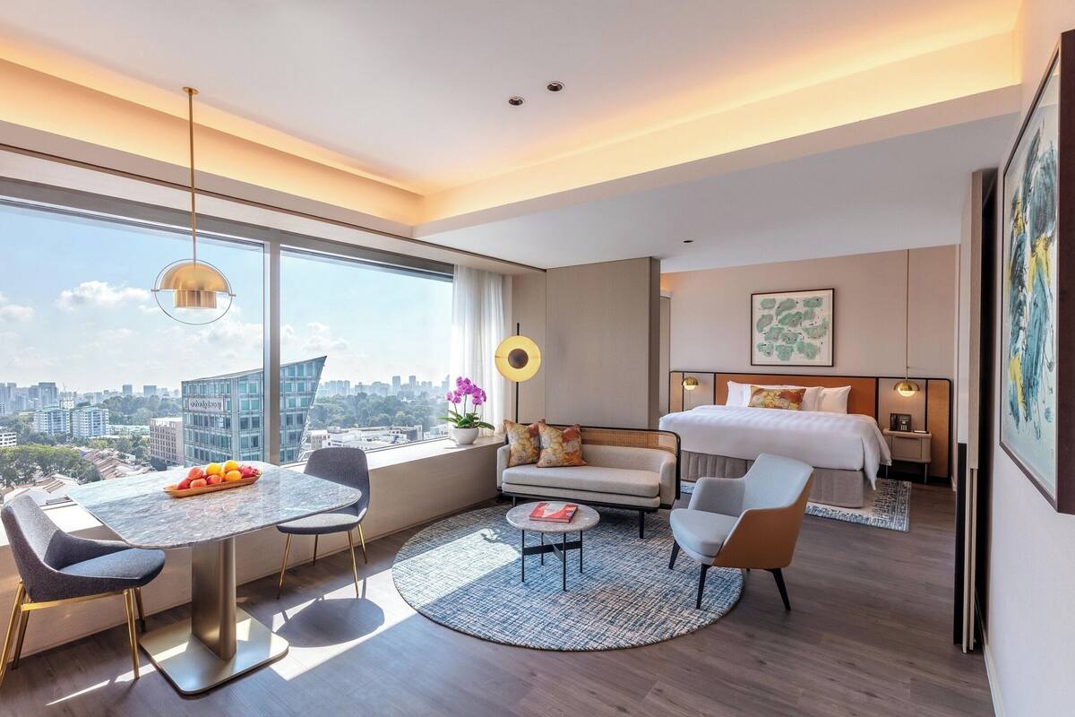 A modern hotel room with a large window, a dining area with a table and chairs, a sofa, a bed, and a small side table.