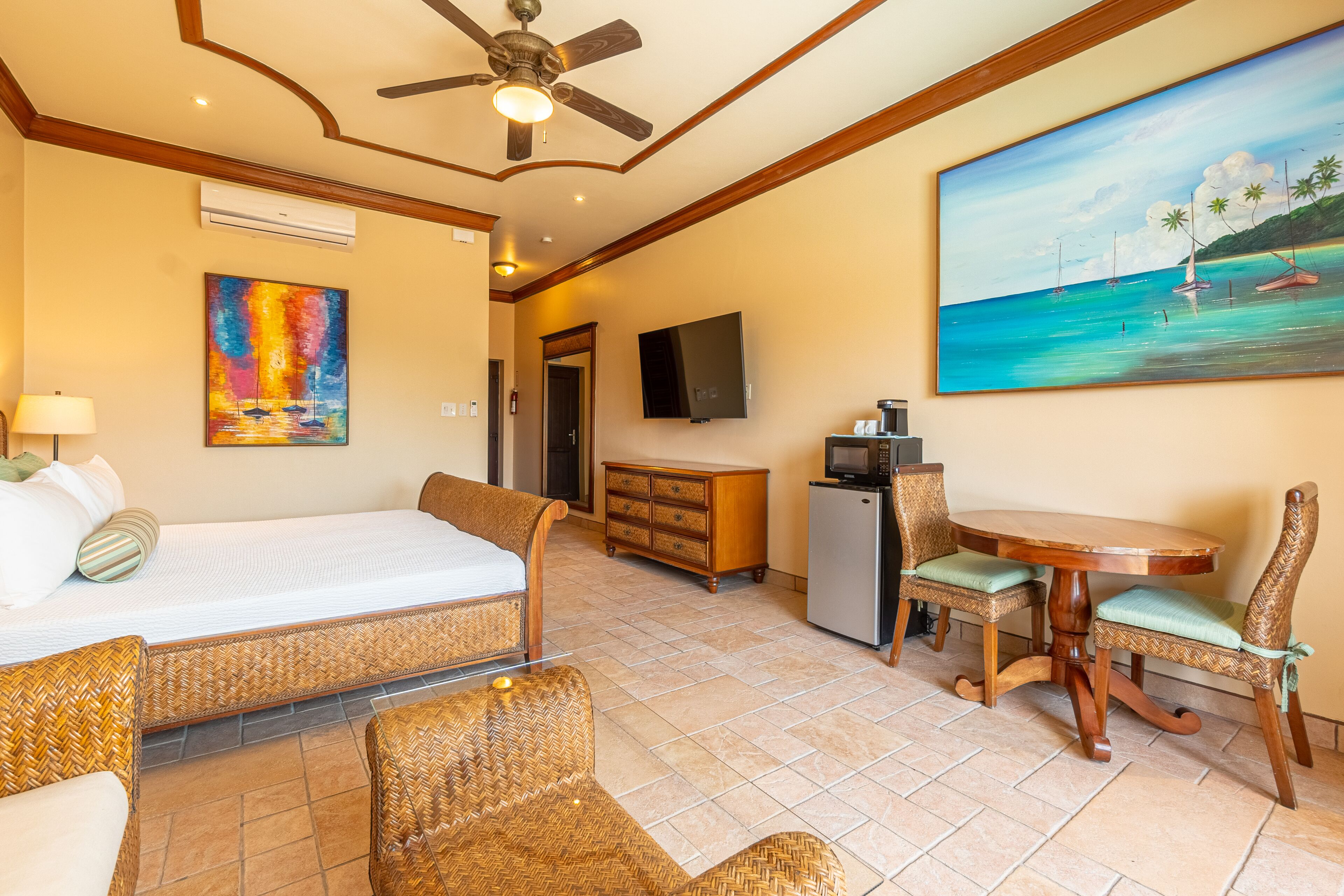 Une chambre d’hôtel comprenant un lit, des meubles en osier, une table à manger avec des chaises, un téléviseur et un tableau représentant une scène de plage.