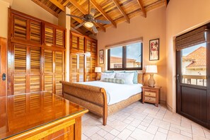 A bedroom with a wooden bed, a glass desk, a ceiling fan, and a window with a view of buildings.