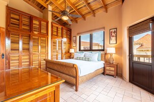 Un dormitorio con una cama de madera, un escritorio de vidrio, un ventilador de techo y una ventana con vistas a edificios.