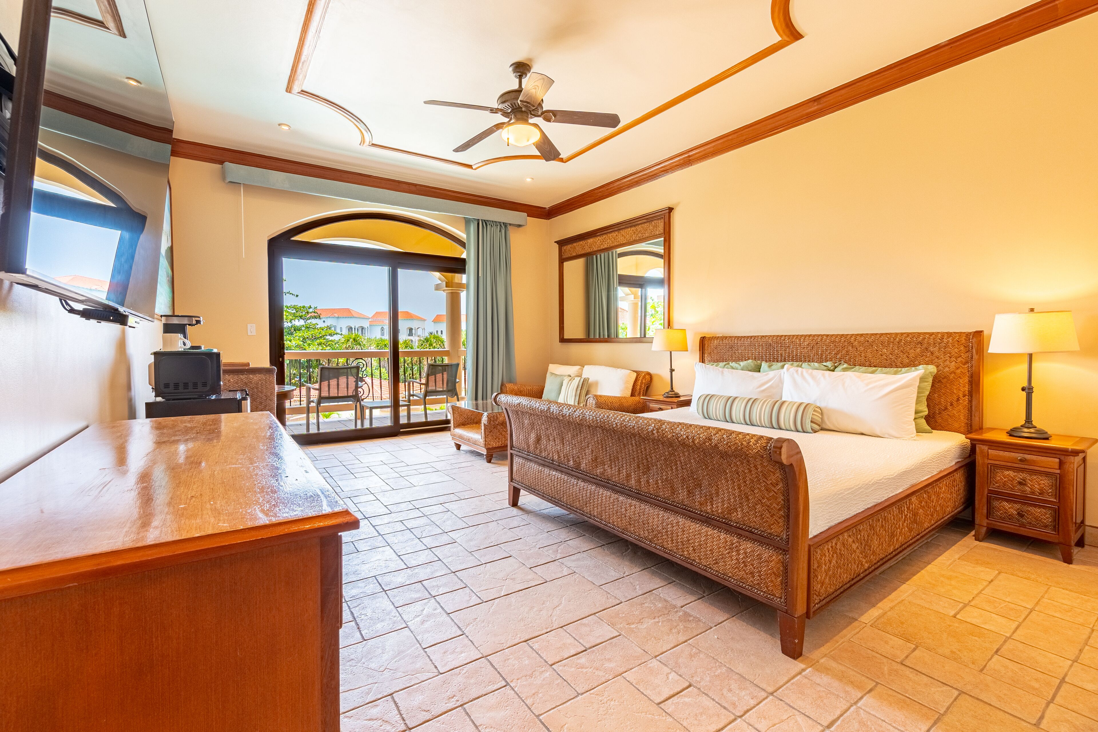 A hotel room with a bed, a desk, a ceiling fan, a balcony with a view, and a large mirror.