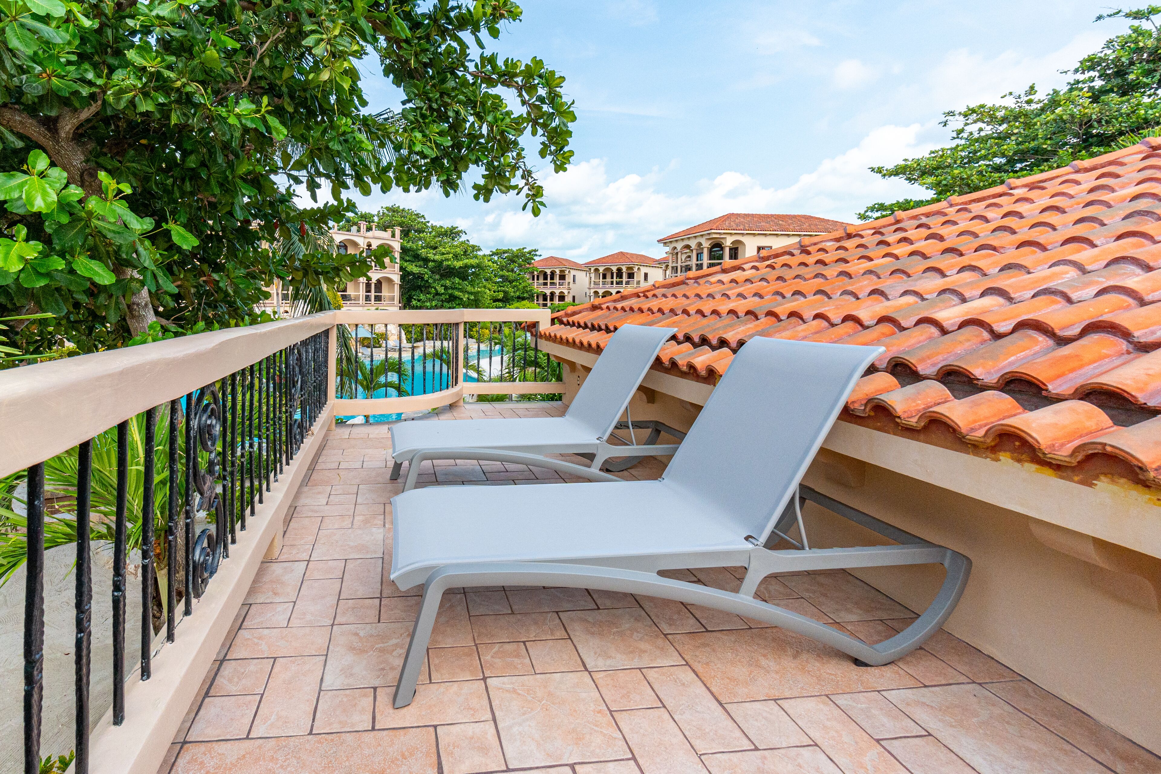 A balcony with two white lounge chairs, a swimming pool, and a tiled roof.