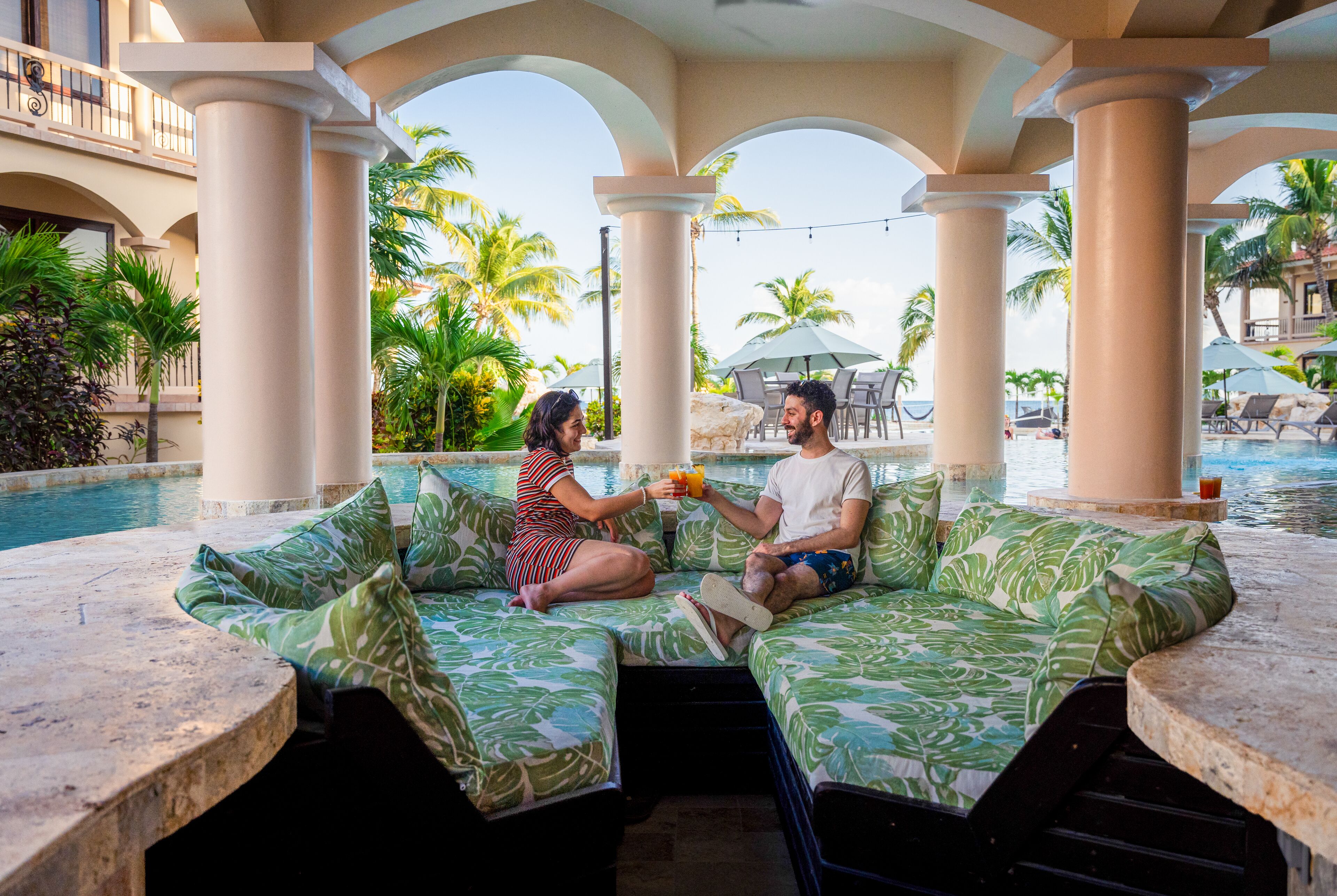 A couple sitting on a poolside sofa with a view of a pool and palm trees.
