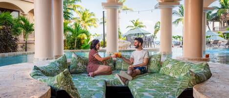 A couple sitting on a poolside sofa with a view of a pool and palm trees.