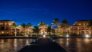 Un hotel frente al agua con un muelle largo que conduce al edificio, rodeado de palmeras e iluminado con luces nocturnas.