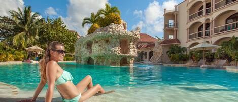 A woman in a bikini by a clear pool with a stone structure and a building with balconies in the background.
