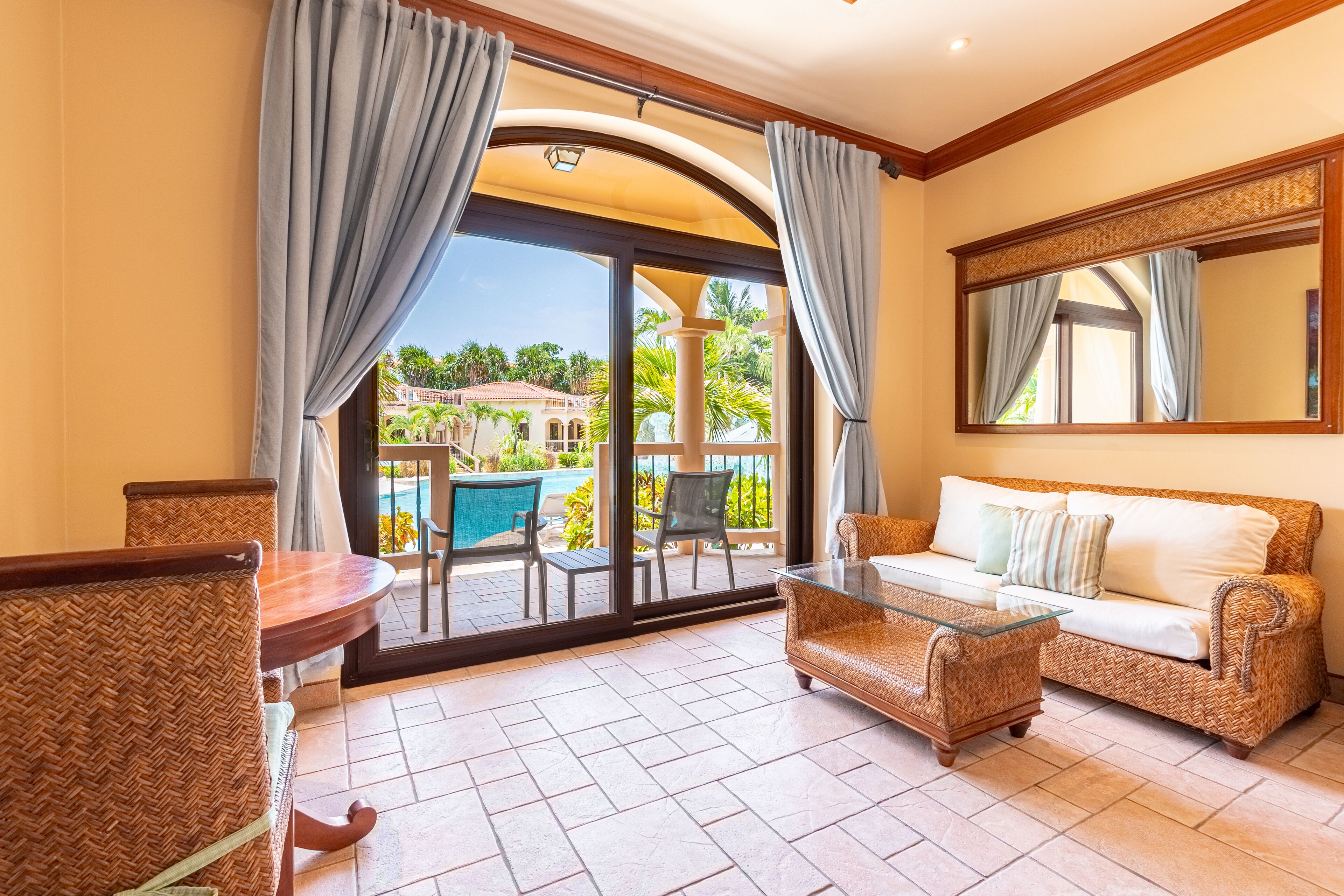 A room with a wicker sofa, a glass coffee table, and a dining table with chairs. There is a large sliding glass door leading to an outdoor area with a pool and palm trees.
