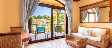 A room with a wicker sofa, a glass coffee table, and a dining table with chairs. There is a large sliding glass door leading to an outdoor area with a pool and palm trees.