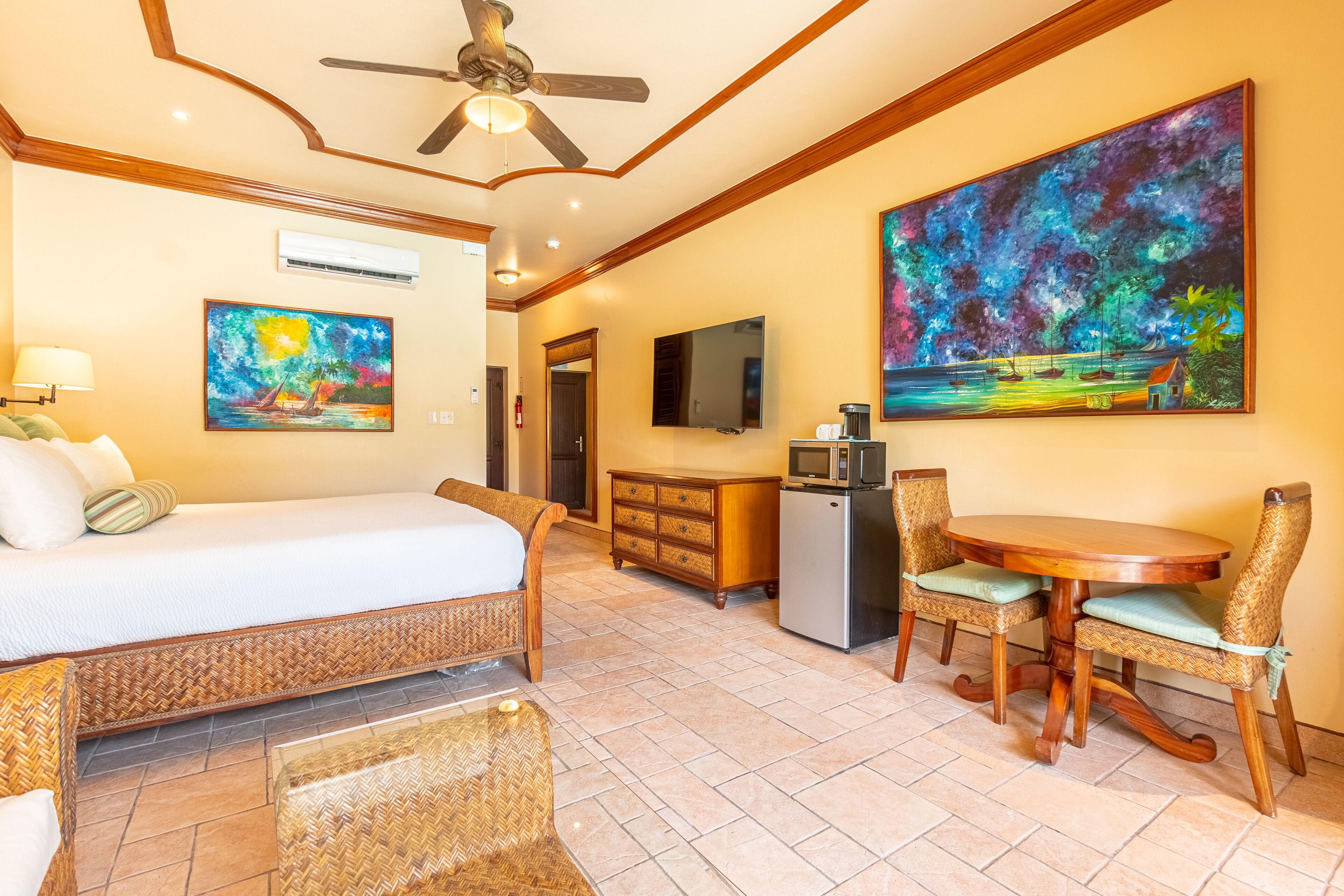 Una habitación de hotel con cama, una mesa de comedor con sillas, ventilador de techo y obras de arte en las paredes.