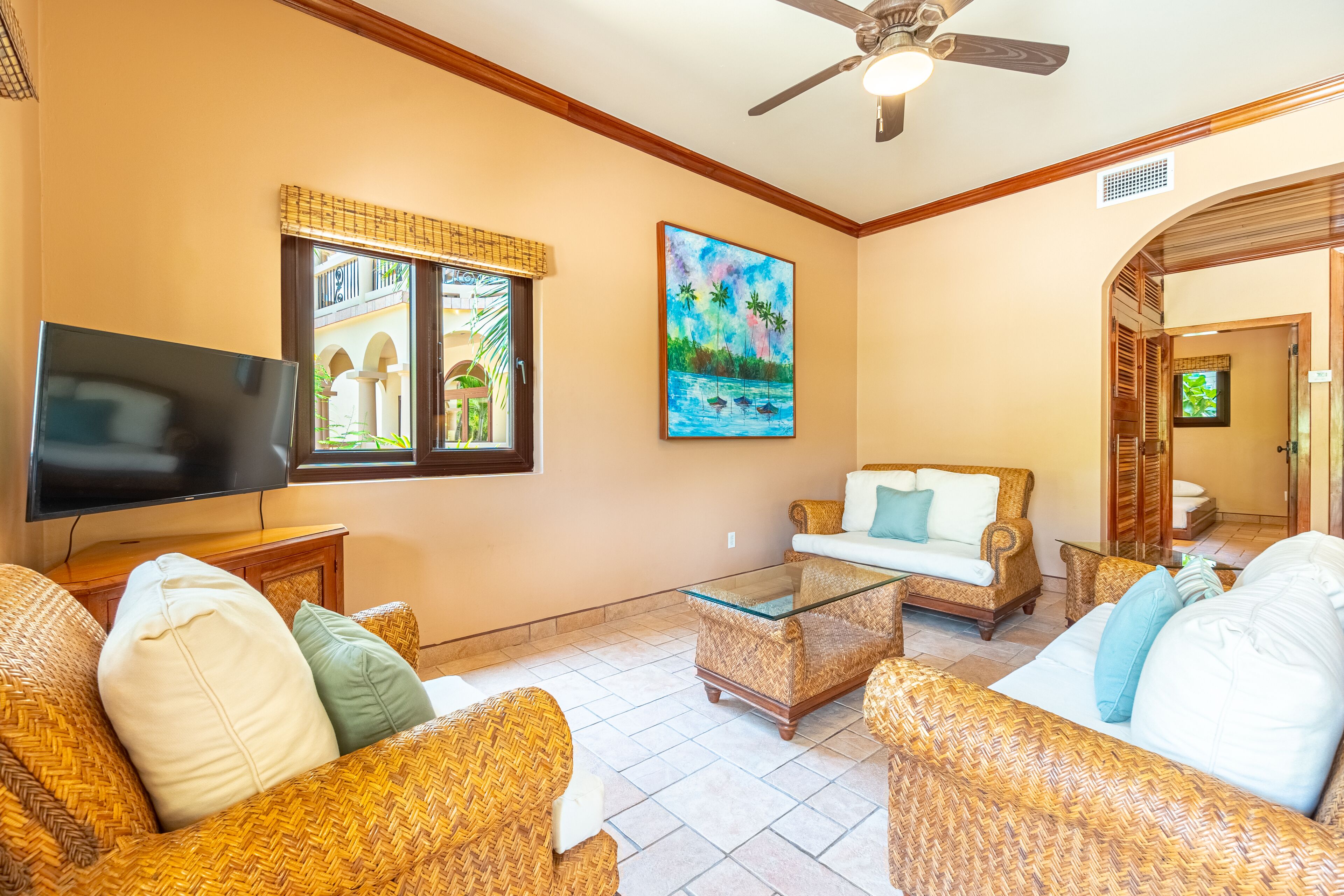 A living room with wicker furniture, a flat-screen TV, and a painting of a tropical scene.