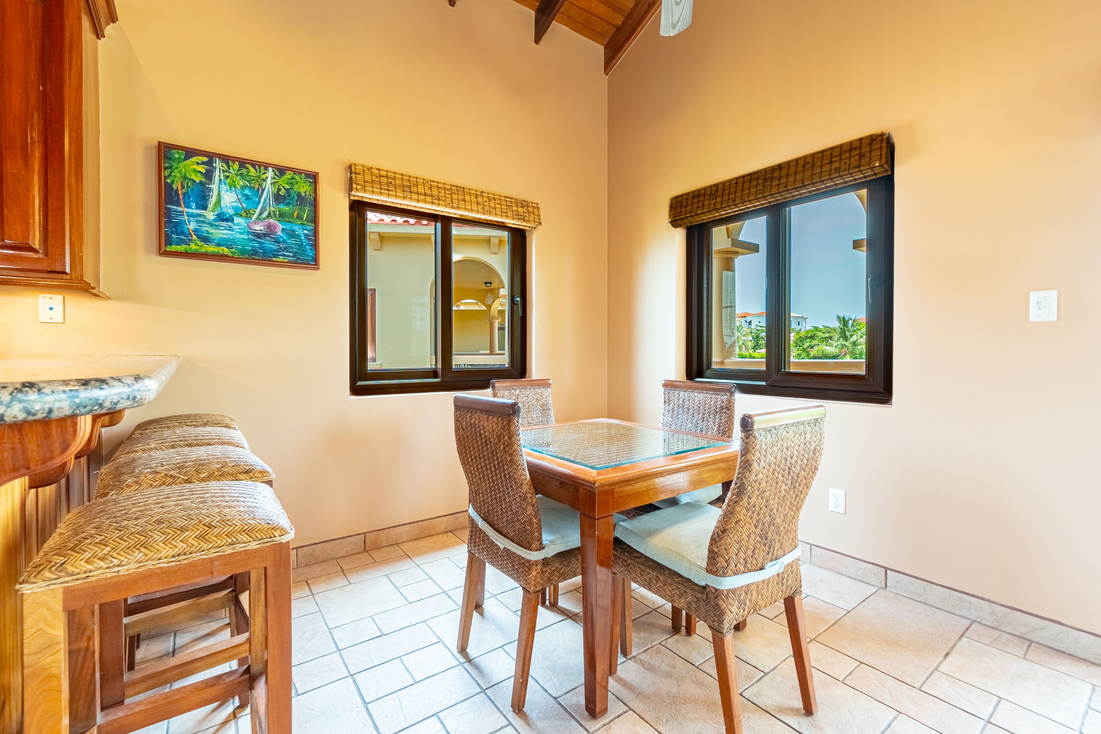 A dining area with a wooden table and wicker chairs, a painting of a tropical scene, and large windows with blinds.