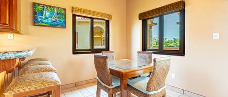 A dining area with a wooden table and wicker chairs, a painting of a tropical scene, and large windows with blinds.