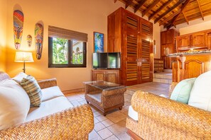 A living room with wicker furniture, a glass coffee table, a TV, wooden cabinets, and a large window with a view of palm trees.