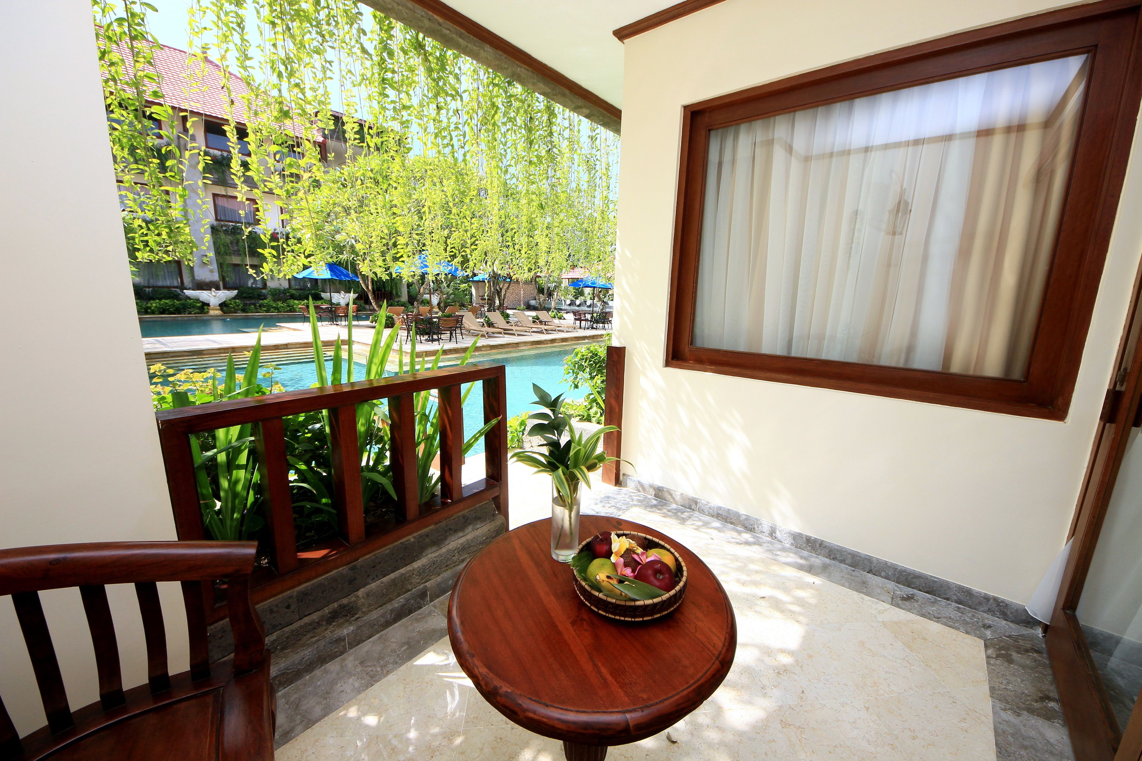 A balcony with a wooden chair, a small table with a fruit bowl, a potted plant, and a view of a pool and buildings.