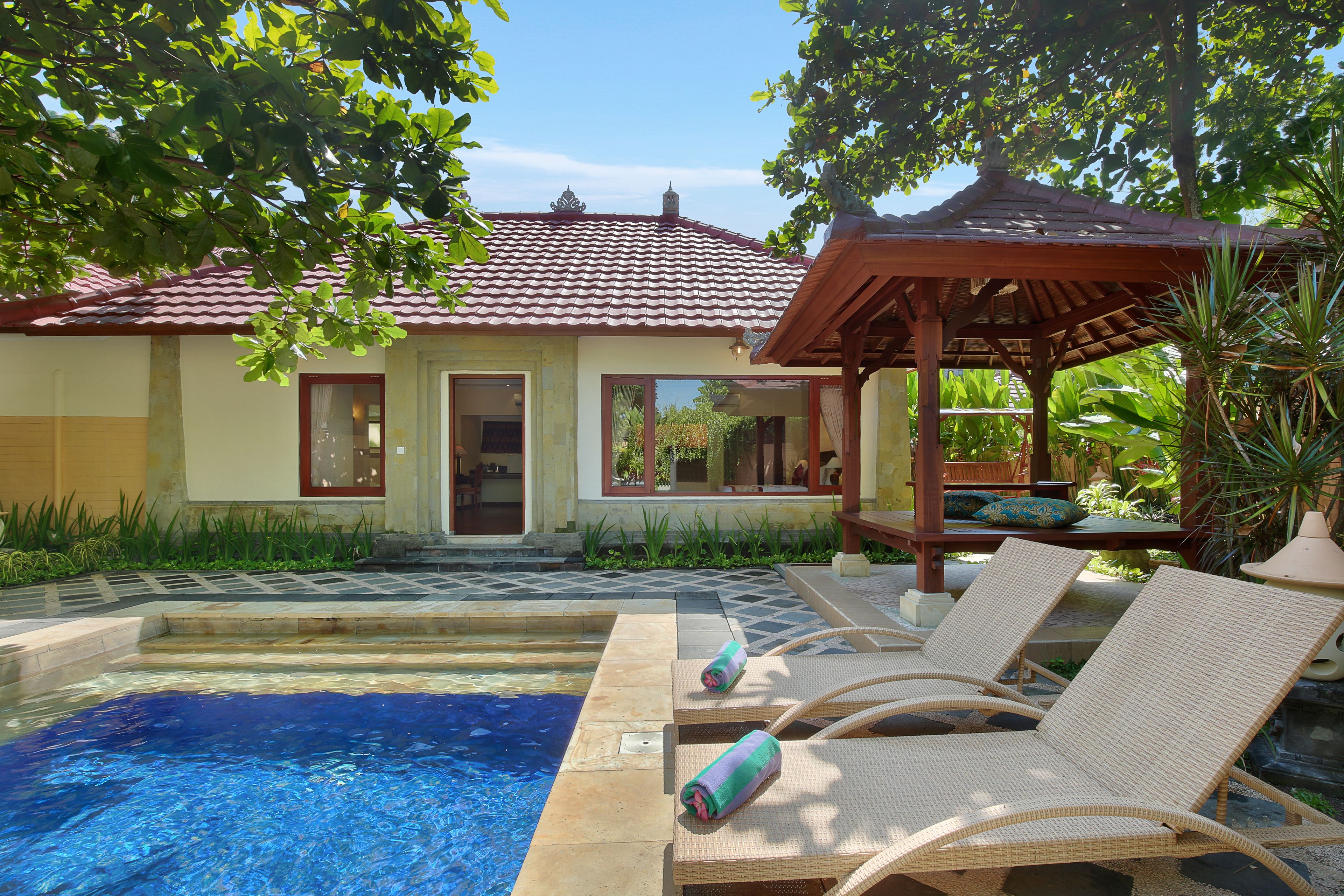 A pool area with lounge chairs and a pergola, surrounded by greenery and a building with a tiled roof.