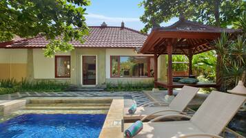 A pool area with lounge chairs and a pergola, surrounded by greenery and a building with a tiled roof.