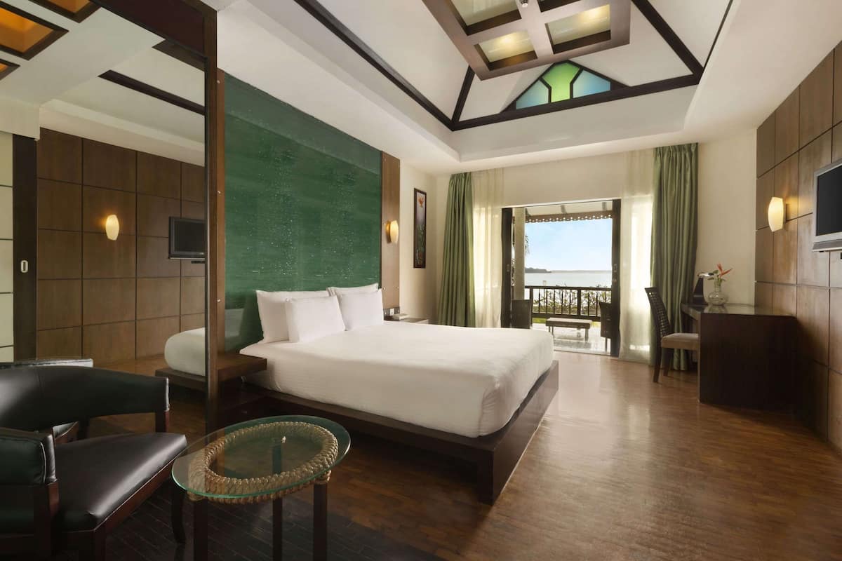 Quarto de hotel com uma cama grande, uma mesa de centro de vidro, uma escrivaninha e vista para o mar.