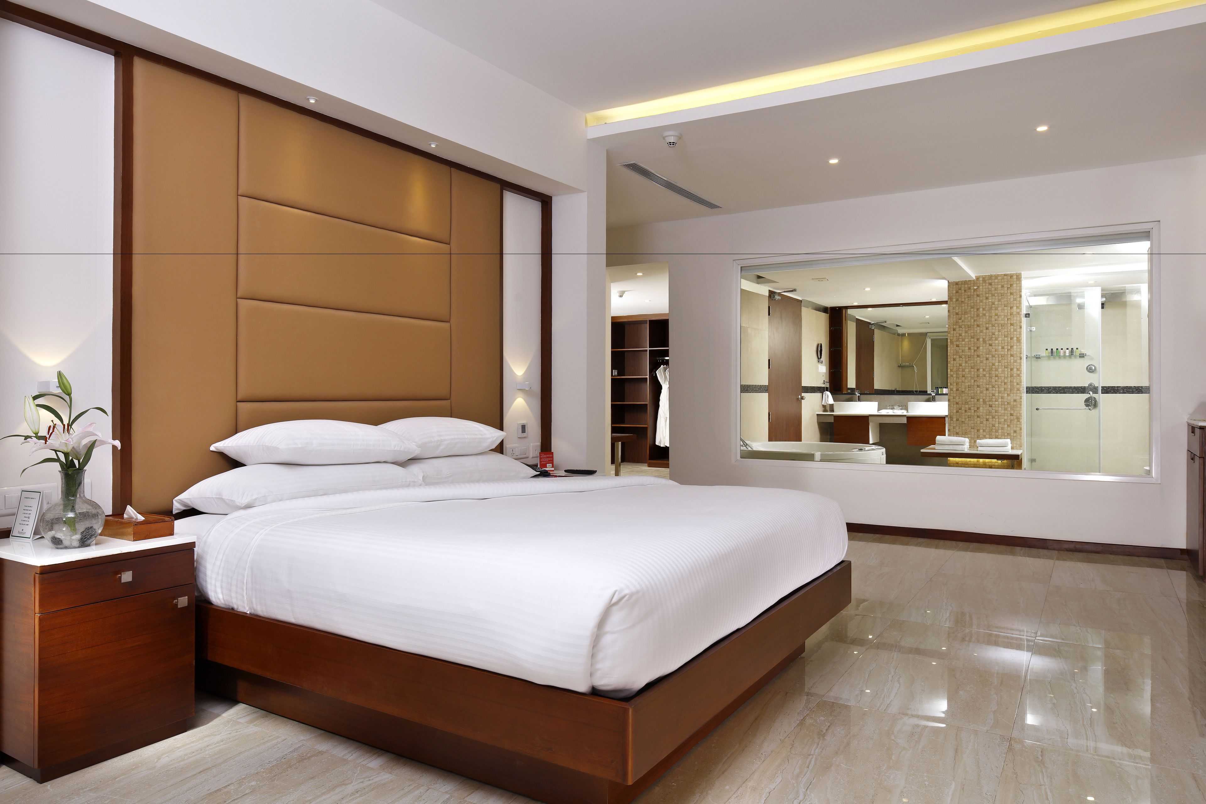 A modern hotel room with a large bed, wooden headboard, bedside tables, a mirror, and a bathroom visible through an open door.