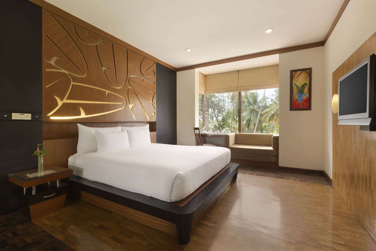 Quarto de hotel moderno com uma cama grande, cabeceira de madeira, televisão e janela com vista para palmeiras.