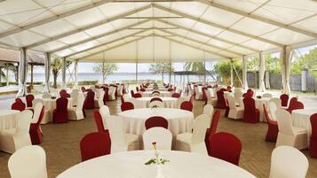 A covered outdoor event space with round tables and chairs, a view of the sea, and a brick floor.