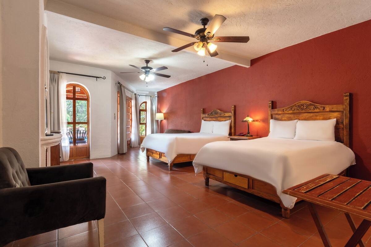 A hotel room with two beds, a wooden headboard, a ceiling fan, and a view of the outdoors through a window.