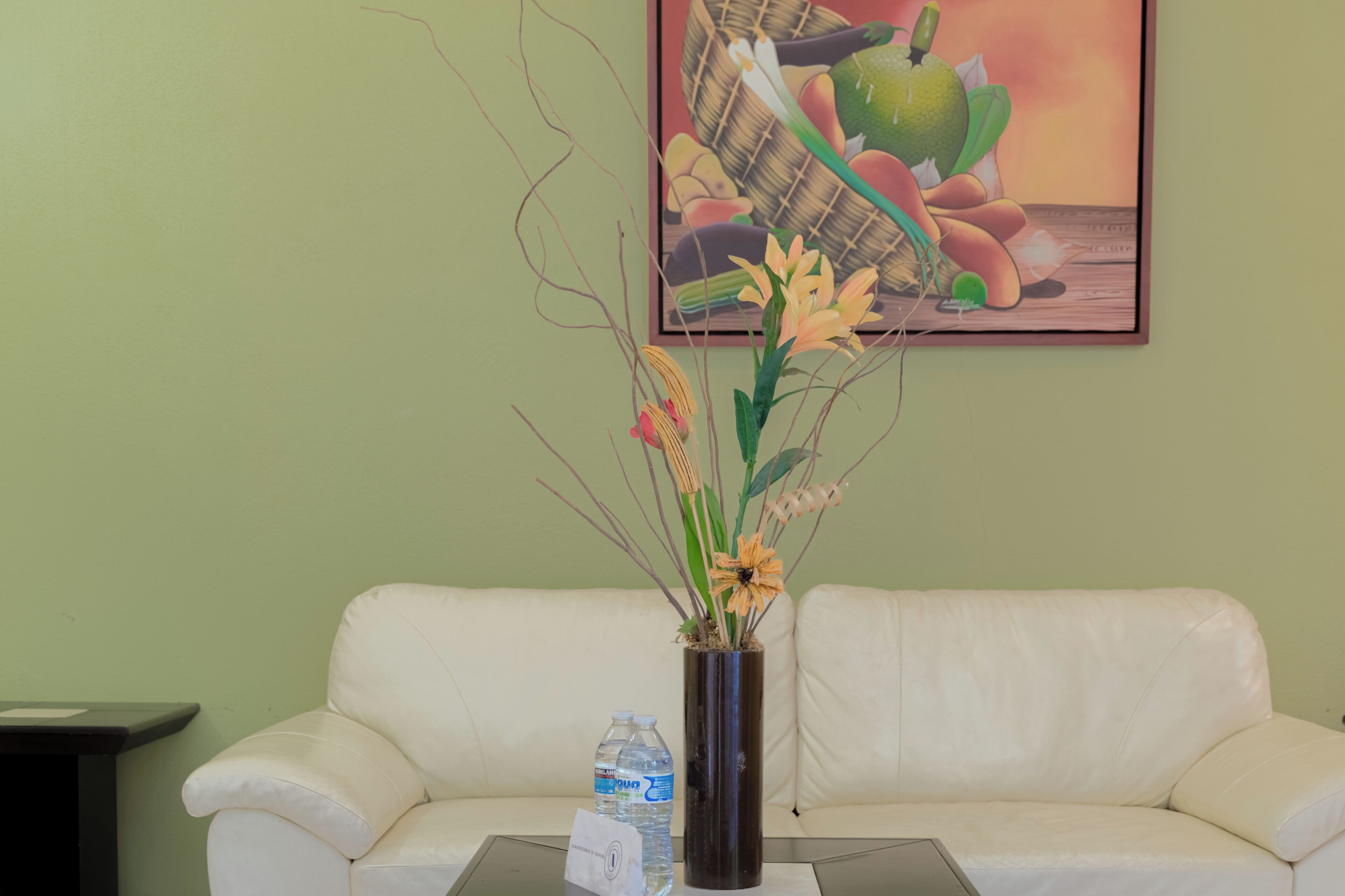 A room with a white leather sofa, a glass coffee table, a vase with flowers, a water bottle, and a painting on the wall.