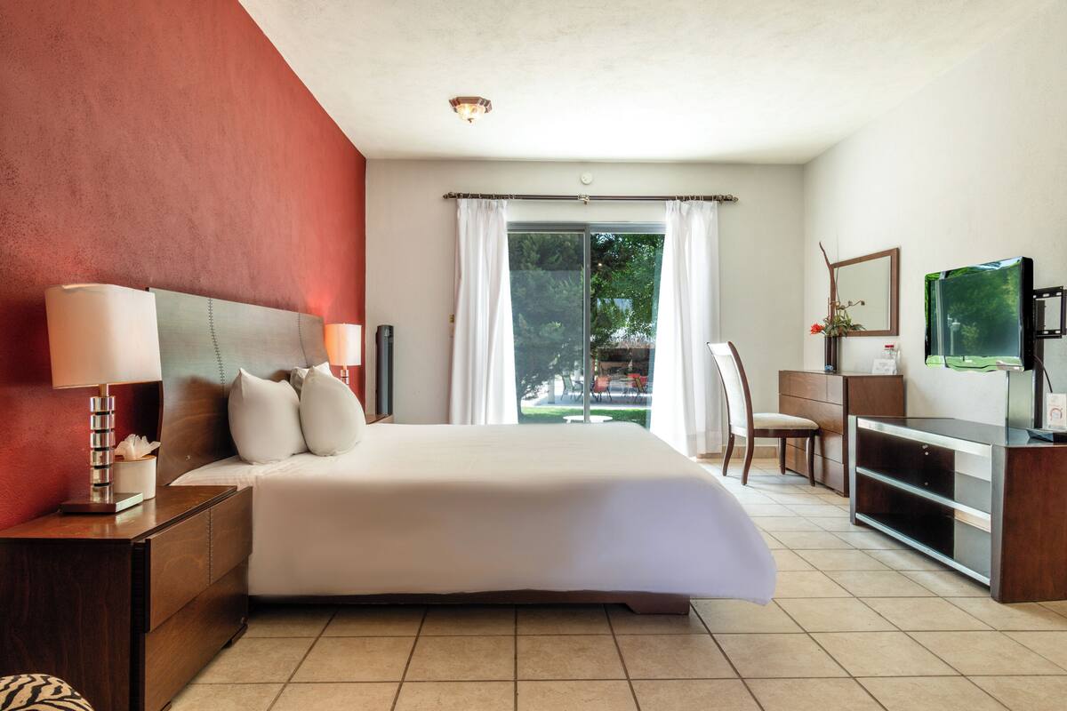 A hotel room with a large bed, a desk with a computer, a chair, a TV, and a window with curtains.