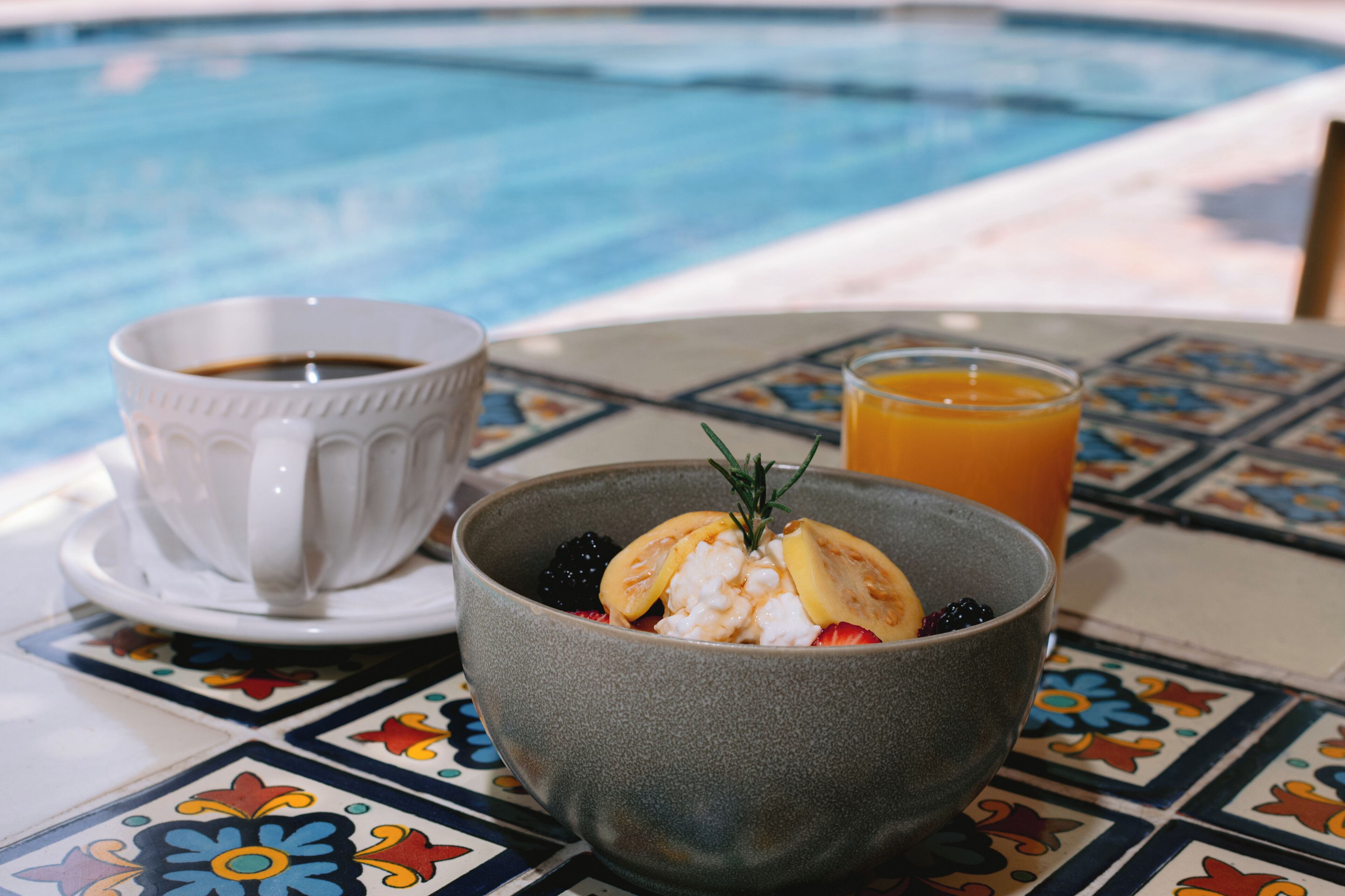 A table with a bowl of breakfast, a cup of coffee, and a glass of orange juice by a pool.