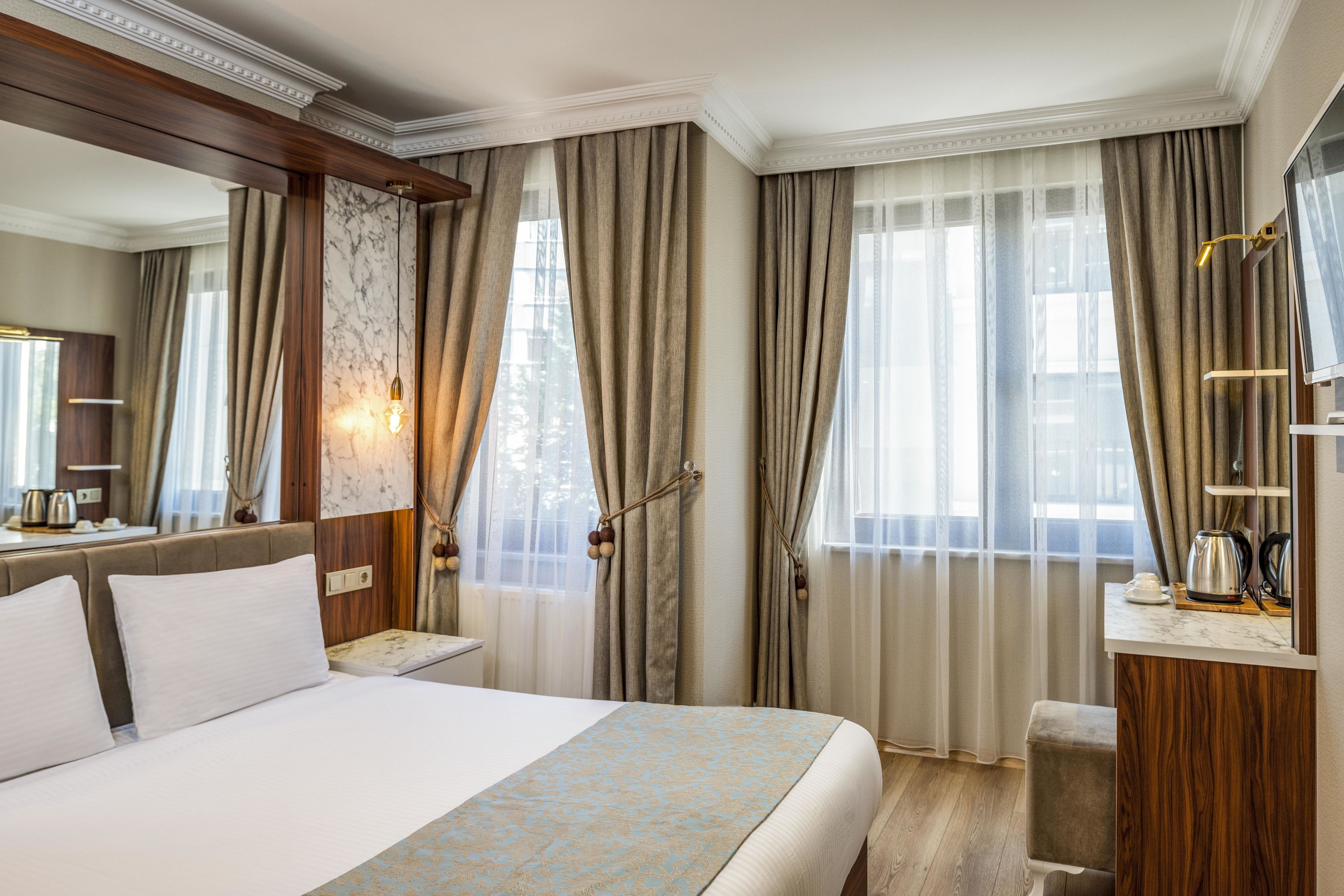 Habitación de hotel con una cama grande, cabecero de madera, escritorio con lámpara, espejo y ventana con cortinas.