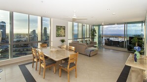 A spacious living area with a dining table, chairs, a sofa, and a TV. Large windows offer a view of the cityscape.
