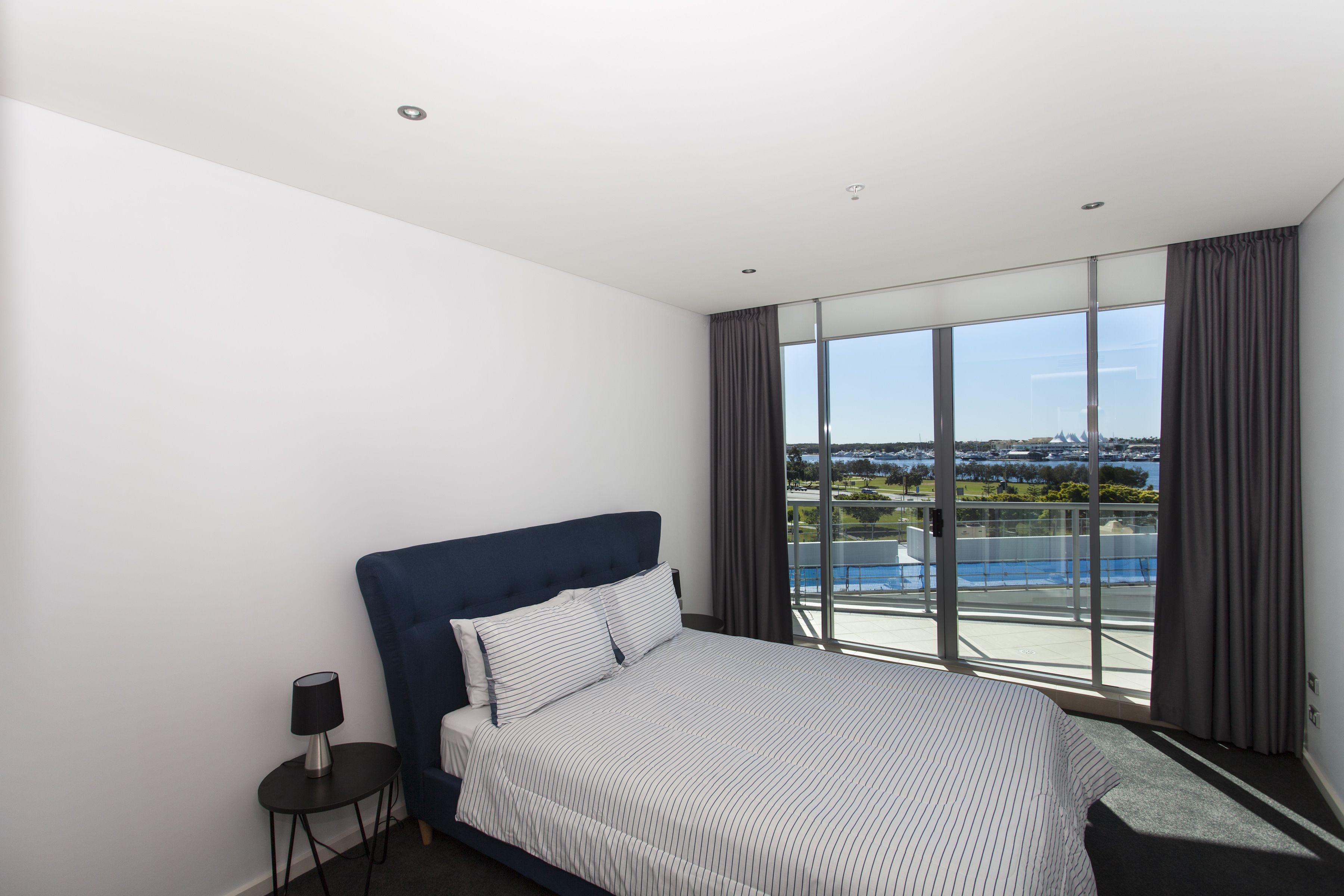 A modern hotel room with a large bed, a small side table, and a view of a cityscape through large windows.