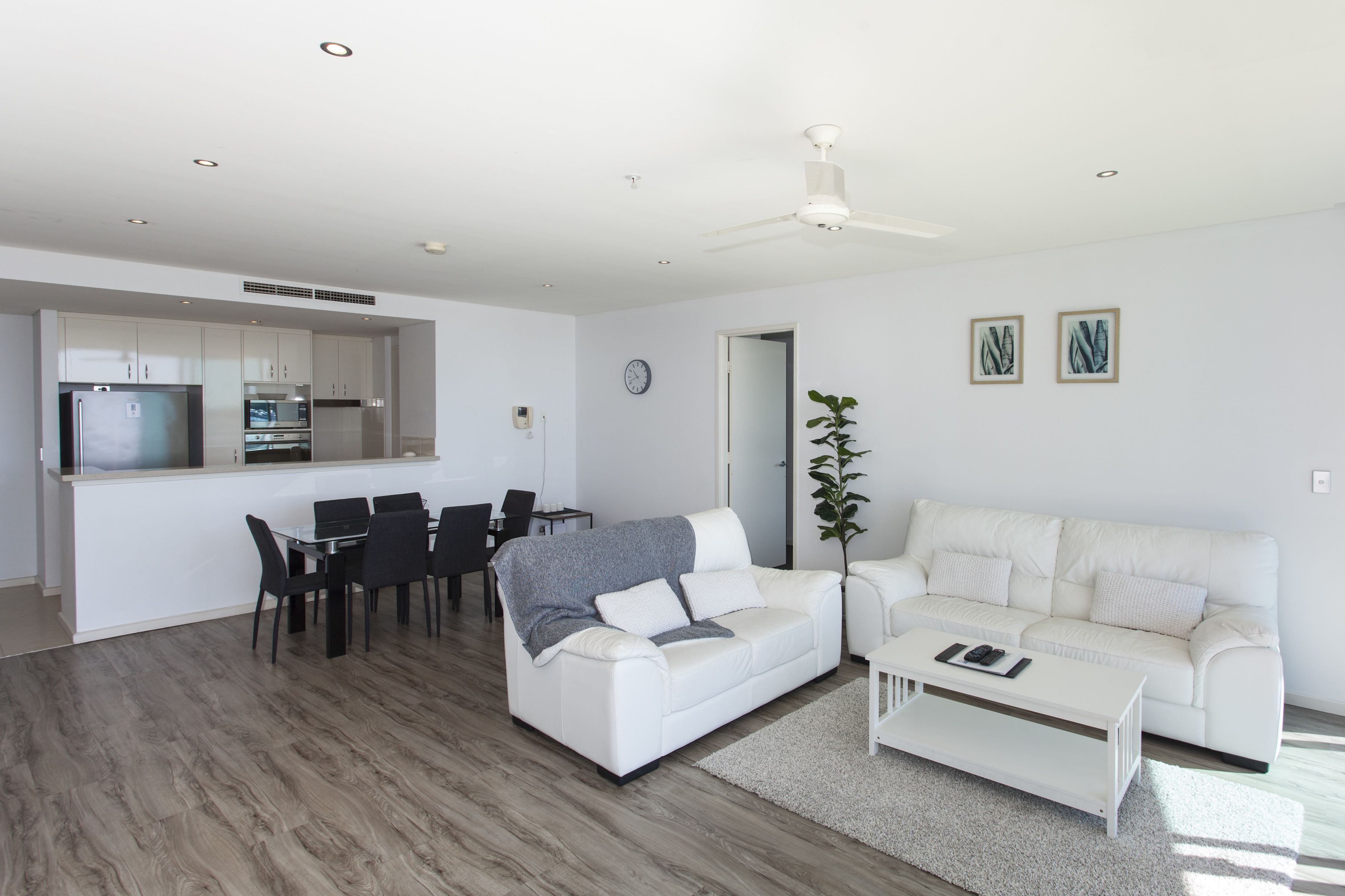 A modern living room with a white sofa, a dining table with chairs, a white coffee table, and a kitchen area with white cabinets and appliances.