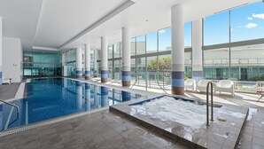An indoor swimming pool with clear glass walls, a hot tub, and a balcony with chairs.