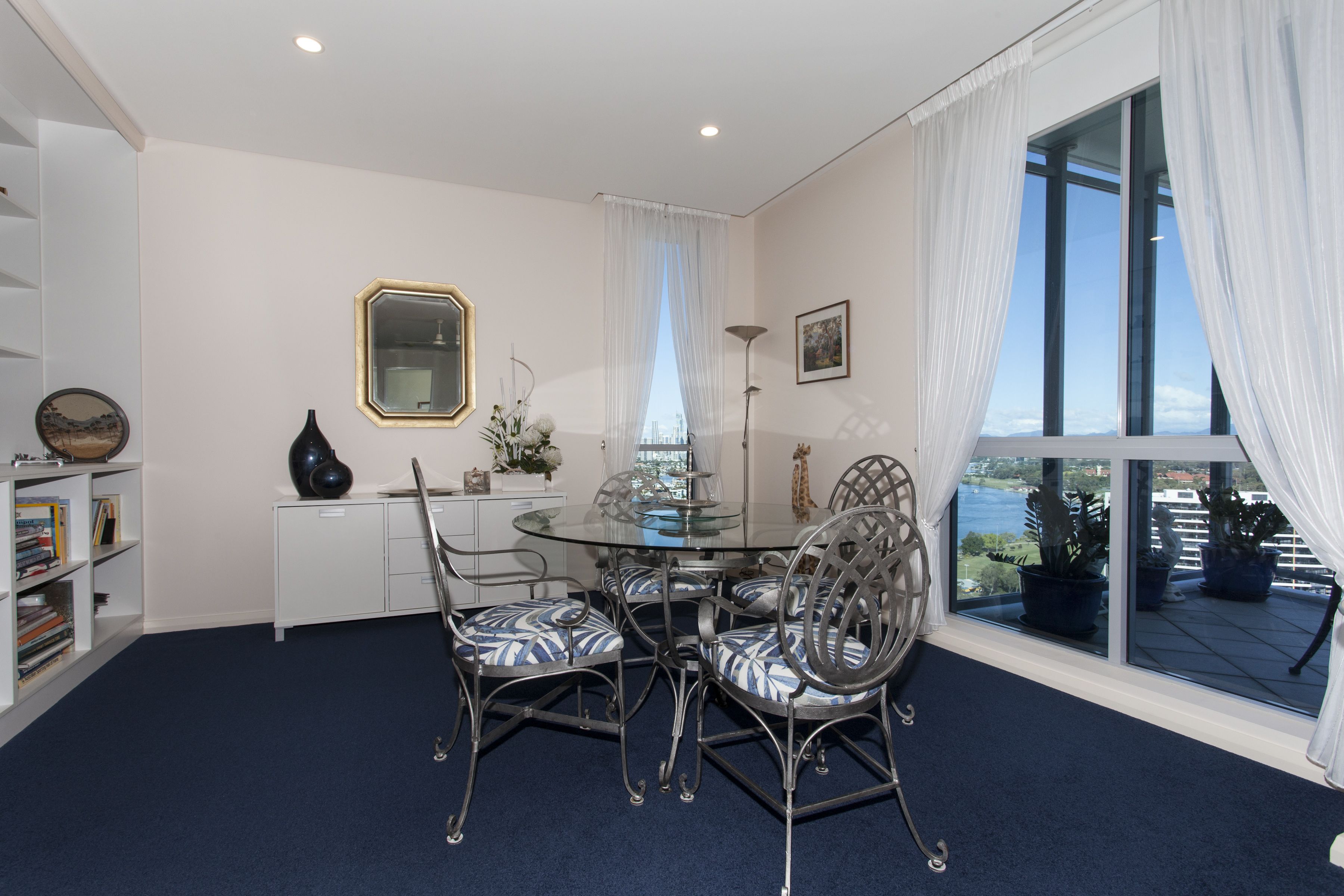 A dining area with a glass table, metal chairs, a white cabinet, and a view of the outdoors through a large window.