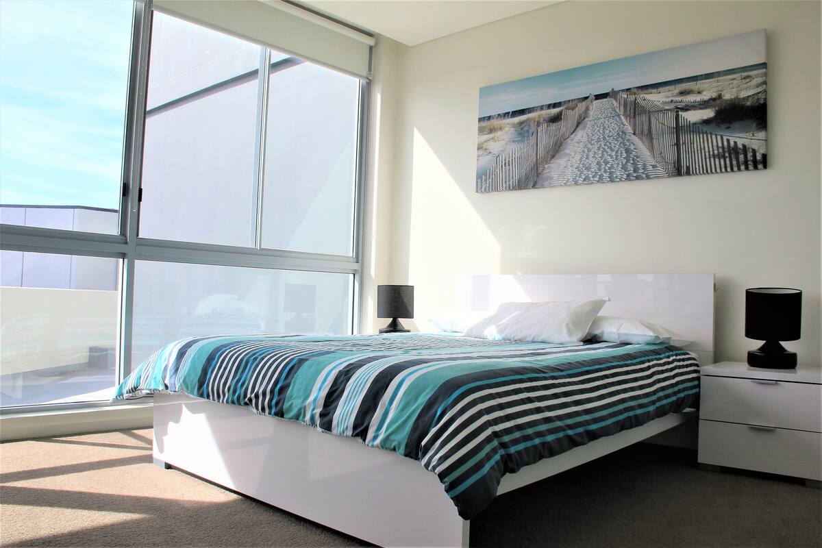 A modern bedroom with a large window, a bed with striped bedding, a nightstand with a lamp, and a painting on the wall.