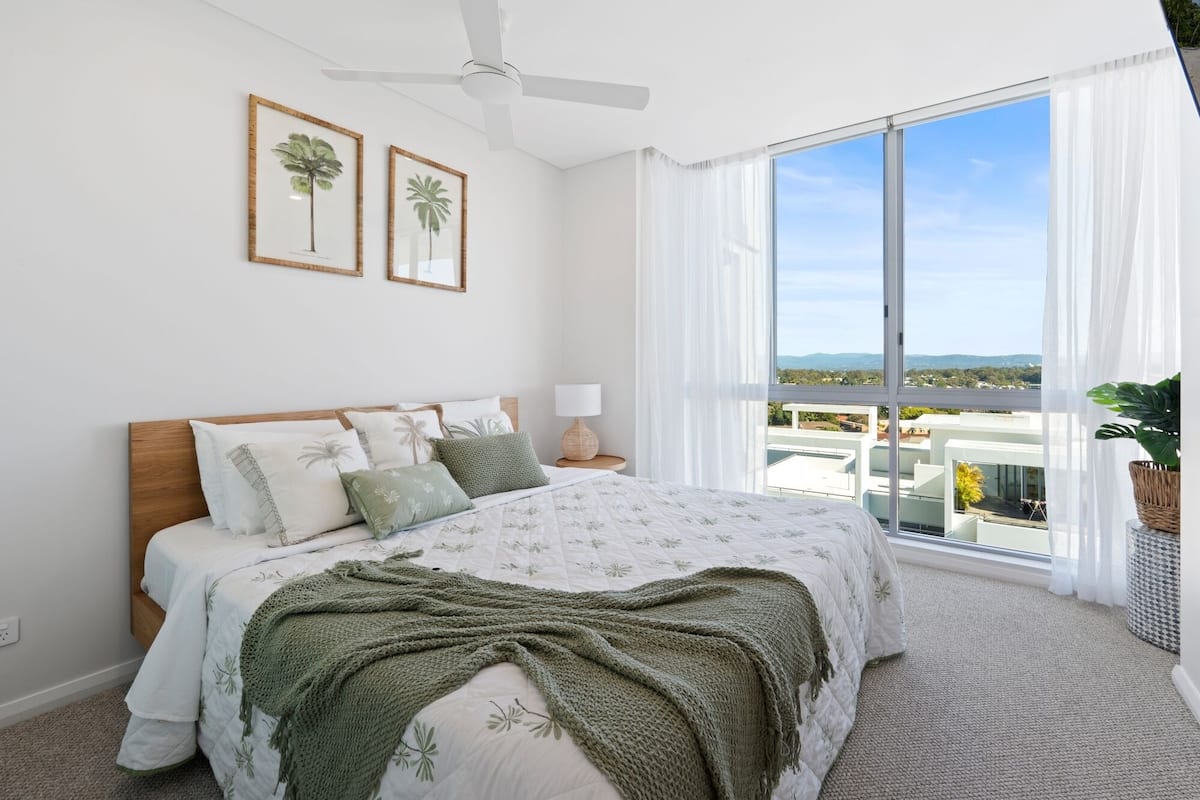 A bedroom with a large bed, a ceiling fan, two framed palm tree pictures, and a view of a cityscape through large windows.