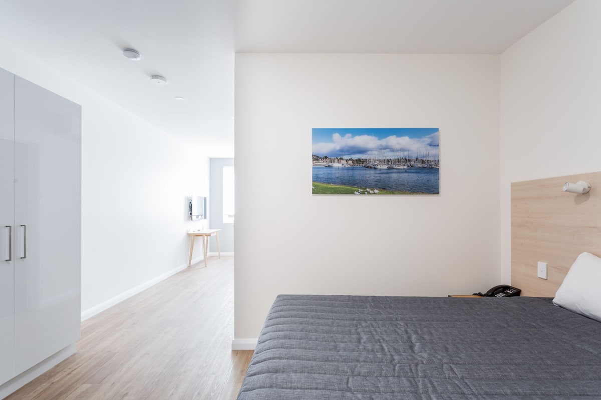 Moderni hotellihuone, jossa on sänky, merimaiseman kuvaava taulu, puinen pääty ja puhelin yöpöydällä.