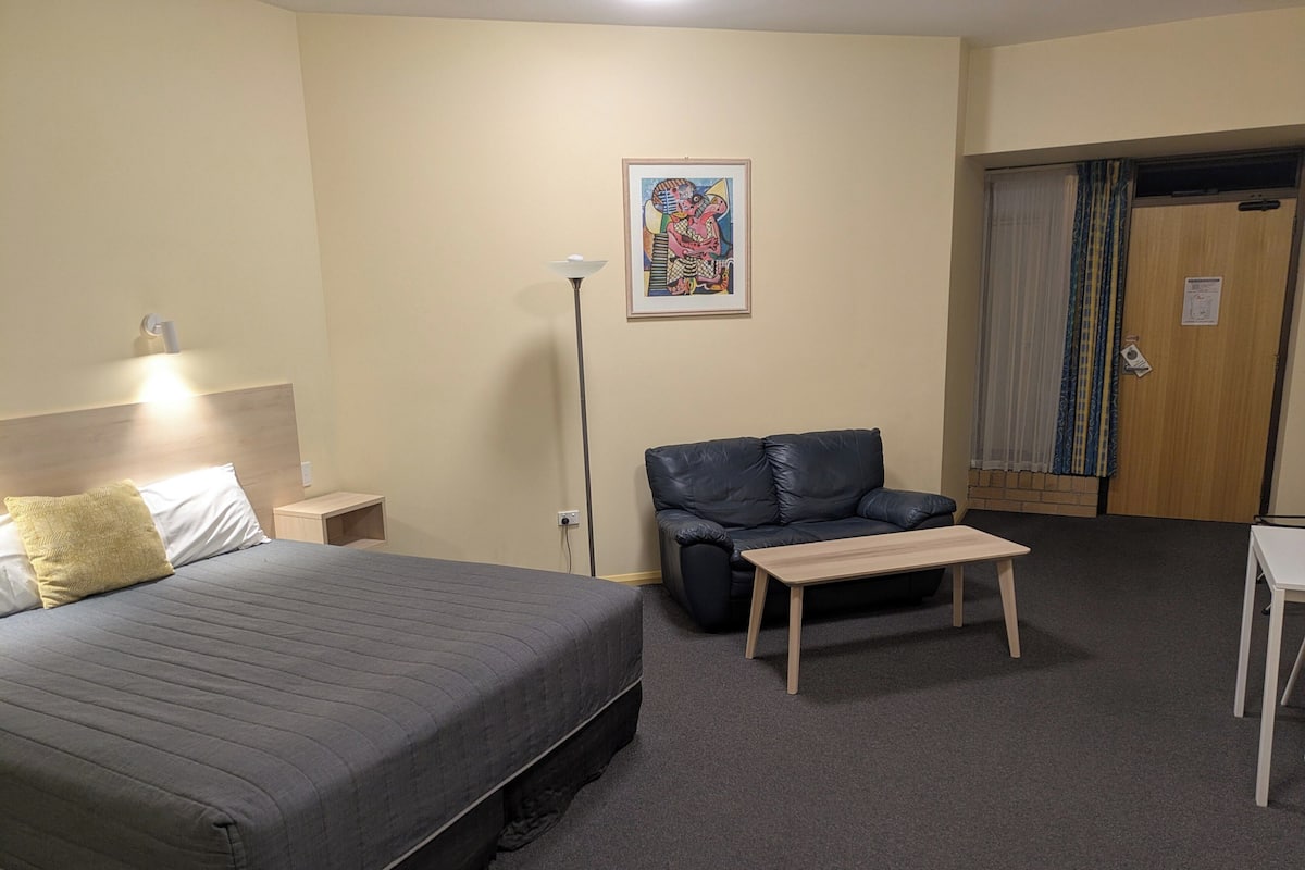 Hotellihuone, jossa on sänky, pieni pöytä, sohva ja taulu seinällä.