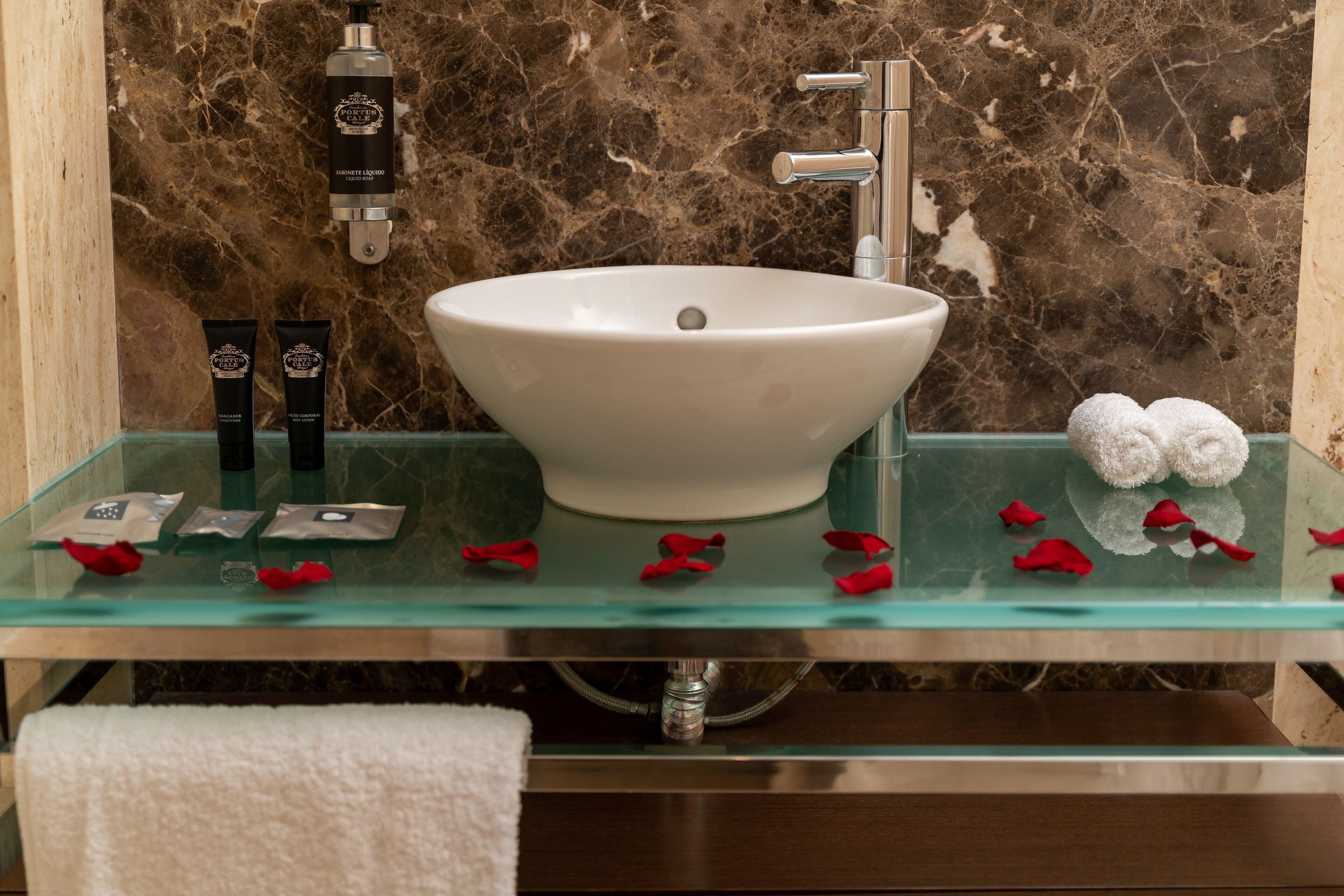 A bathroom with a marble wall, a white vessel sink, and a glass shelf with toiletries and rose petals.